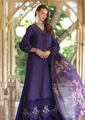 A woman models a deep purple traditional outfit with eyelet embroidery, featuring a long kurta, matching skirt, and a sheer floral dupatta, set against an arched wooden corridor with green foliage in the background.