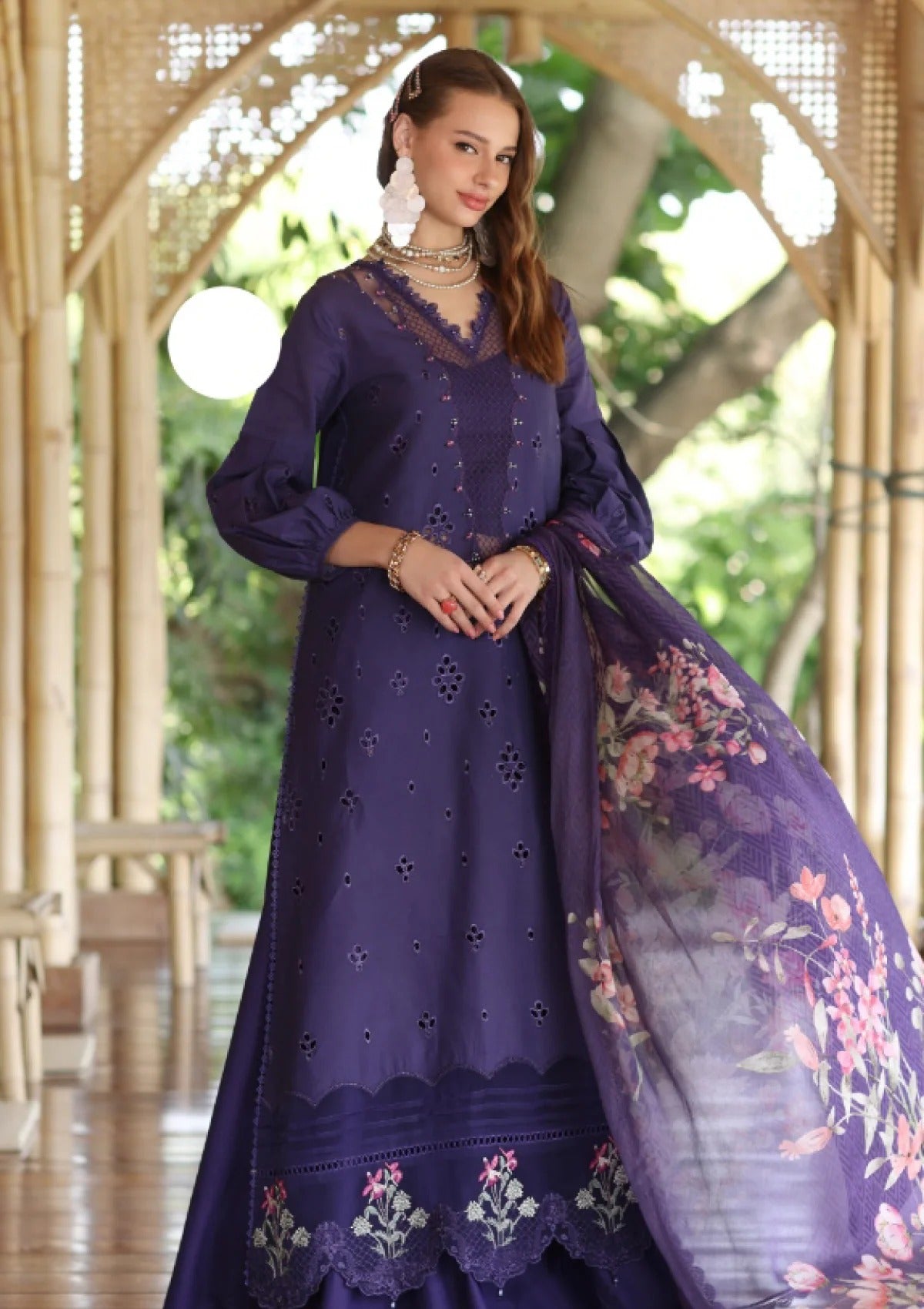 A woman models a deep purple traditional outfit with eyelet embroidery, featuring a long kurta, matching skirt, and a sheer floral dupatta, set against an arched wooden corridor with green foliage in the background.