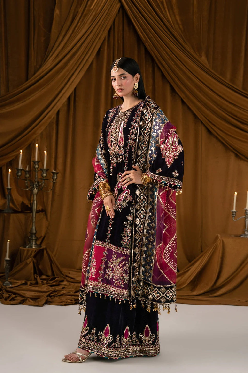 A woman models an ornate traditional outfit with a dark navy base and rich magenta, gold, and beige embroidery, set against a draped brown fabric backdrop with candelabras.