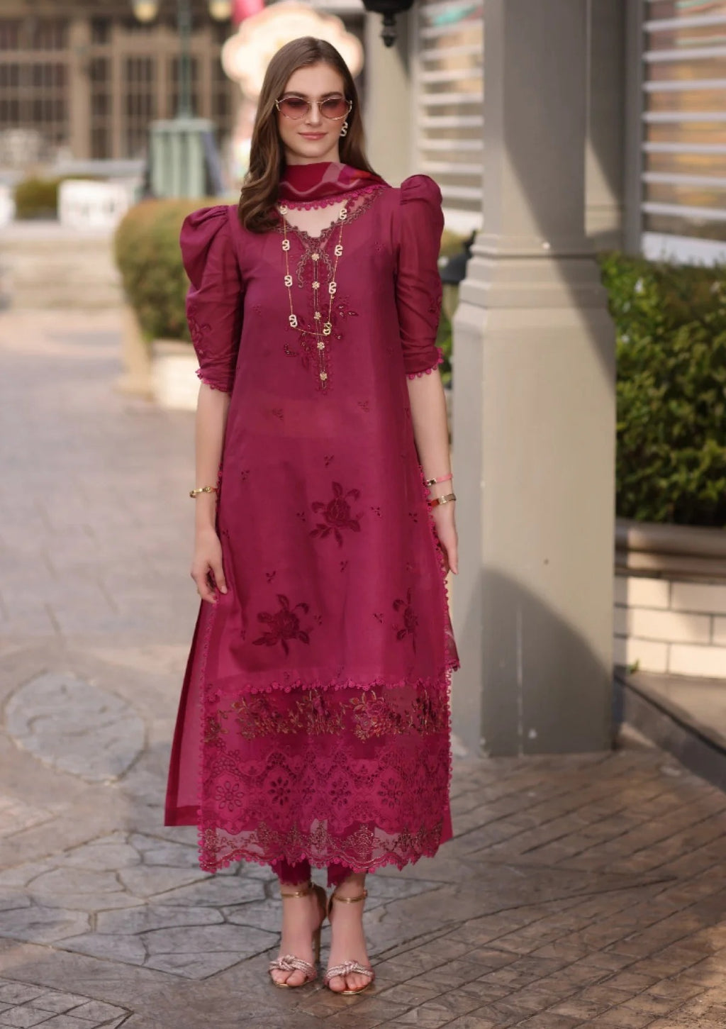 A woman wearing a deep magenta pink embroidered kurta dress with puff sleeves, lace hem, and gold embellishments, standing outdoors on a paved walkway.