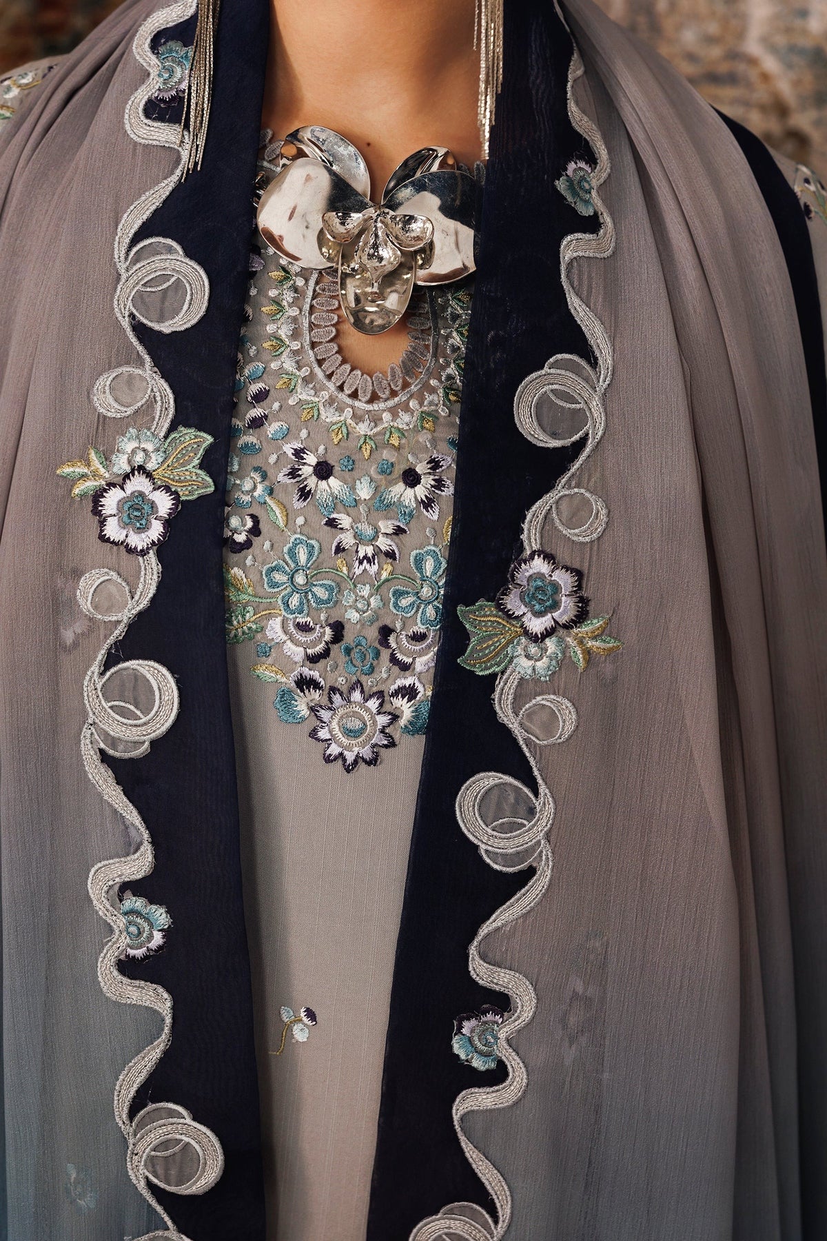 A close-up of traditional South Asian formal wear featuring a taupe-gray embroidered tunic with a large metallic silver floral collar, a dark navy border with silver embroidered swirls and floral motifs, and a matching sheer taupe dupatta.