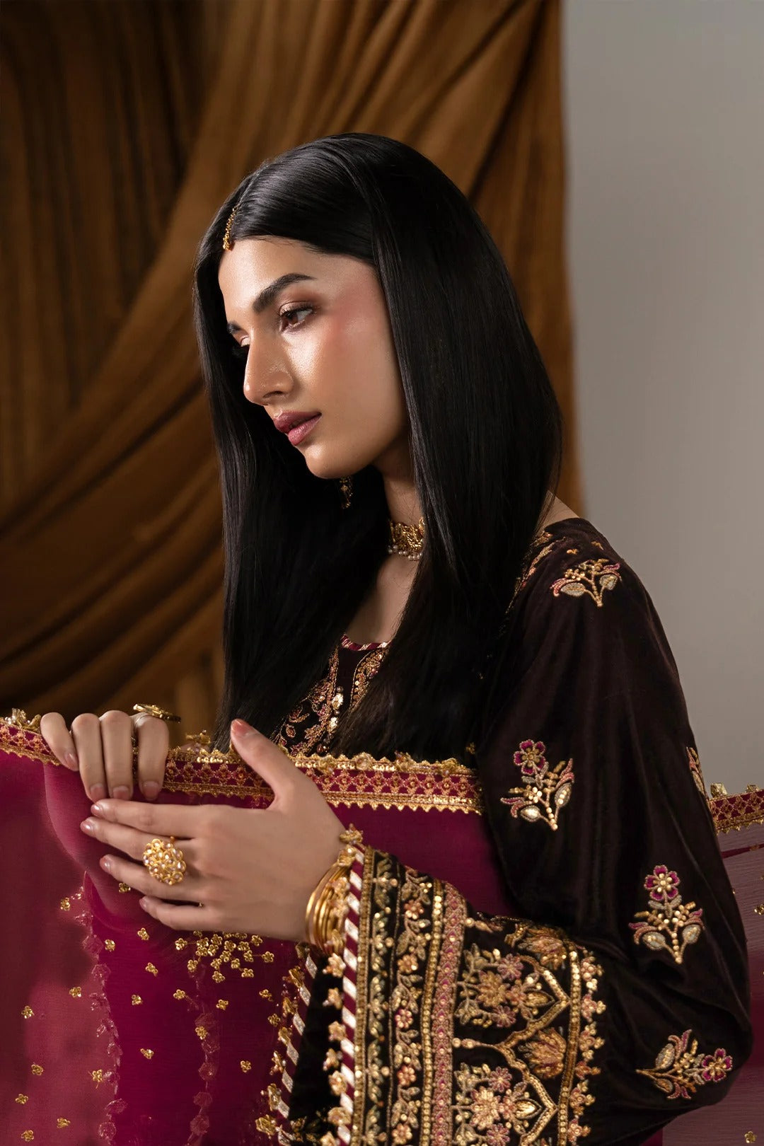 A woman wearing an ornate dark brown and magenta traditional outfit with extensive gold embroidery, holding a matching magenta dupatta. She is styled with gold jewelry against a background of draped brown curtains.