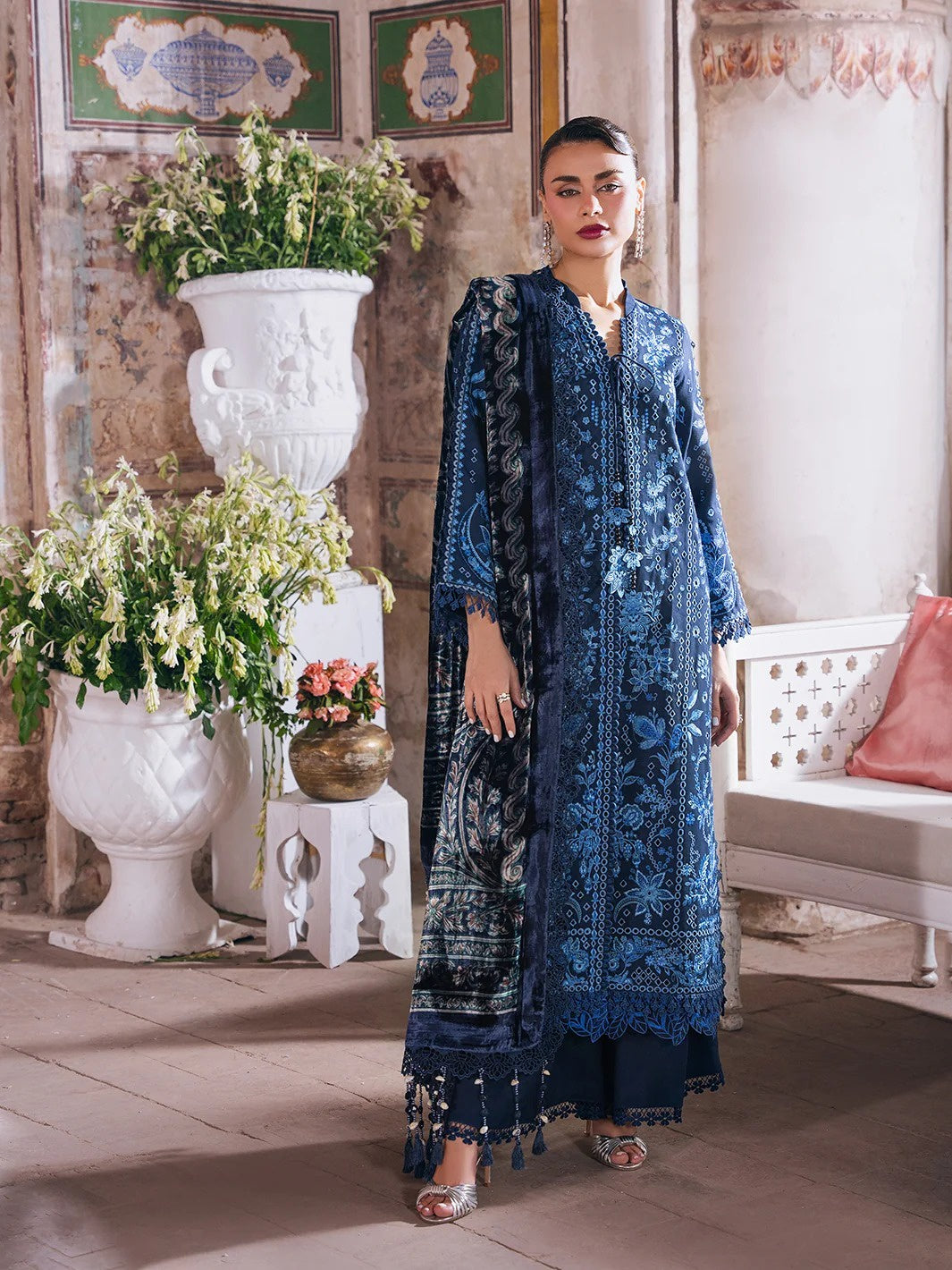 A woman models a traditional blue embroidered salwar kameez set with a matching dupatta in an ornate indoor setting with white planters and a bench.