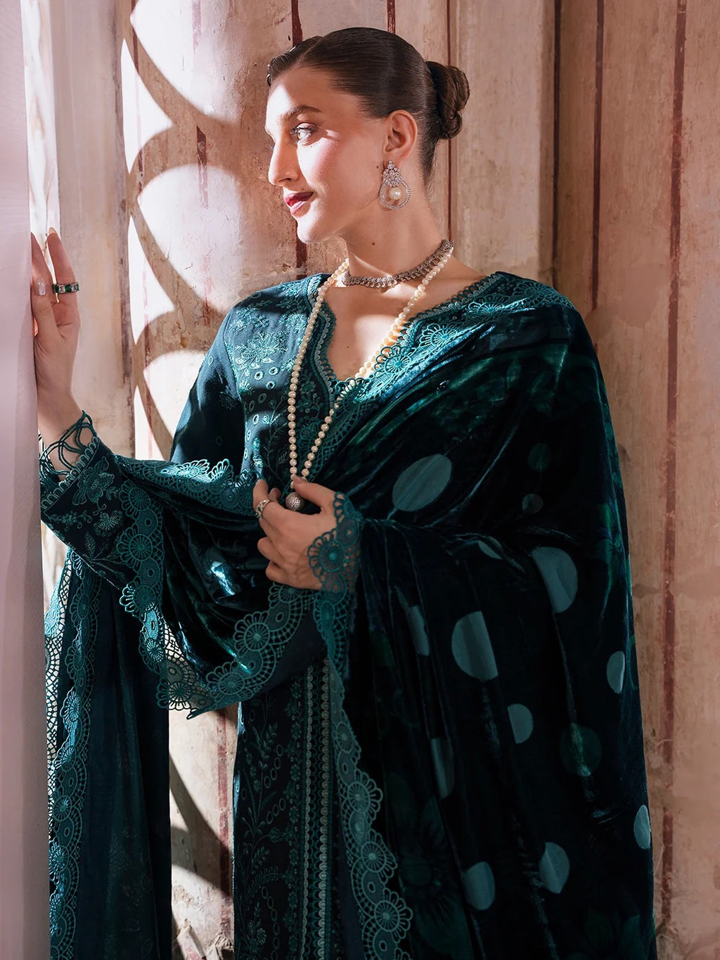 A woman wearing a dark teal velvet and lace traditional outfit with large circular patterns, standing in front of a warm-toned wooden wall with sunlight filtering through.