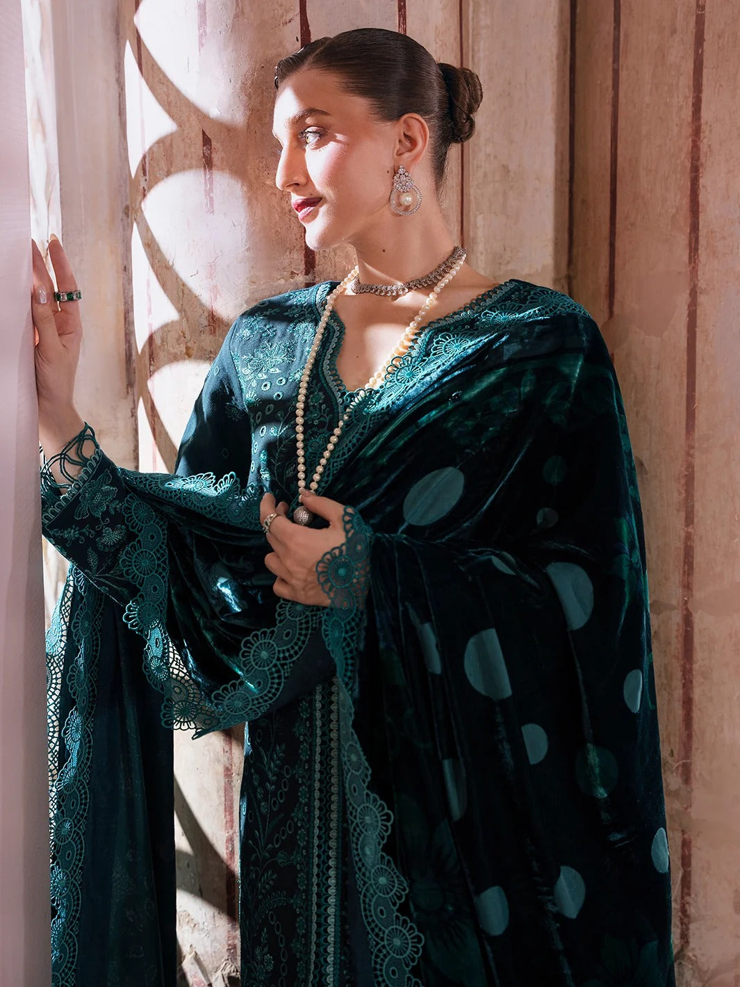 A woman wearing a dark teal velvet and lace traditional outfit with large circular patterns, standing in front of a warm-toned wooden wall with sunlight filtering through.