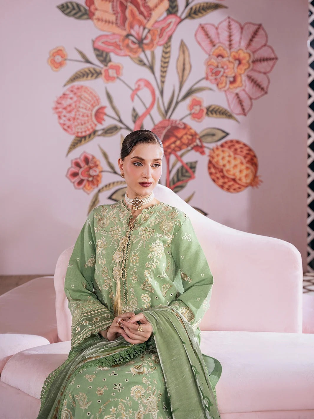 A woman seated on a pale pink sofa wearing a light green traditional embroidered outfit with metallic detailing and a matching dupatta, posed against a pink wall with large floral and pomegranate illustrations.