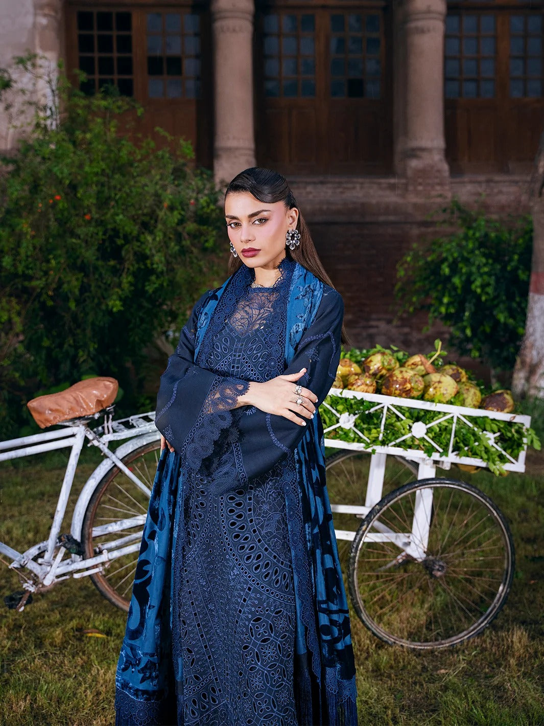 A woman stands outdoors in front of a building and a white bicycle cart, wearing a long dark blue embroidered dress with a matching sheer blue outer layer. The scene includes greenery and rustic architecture in the background.
