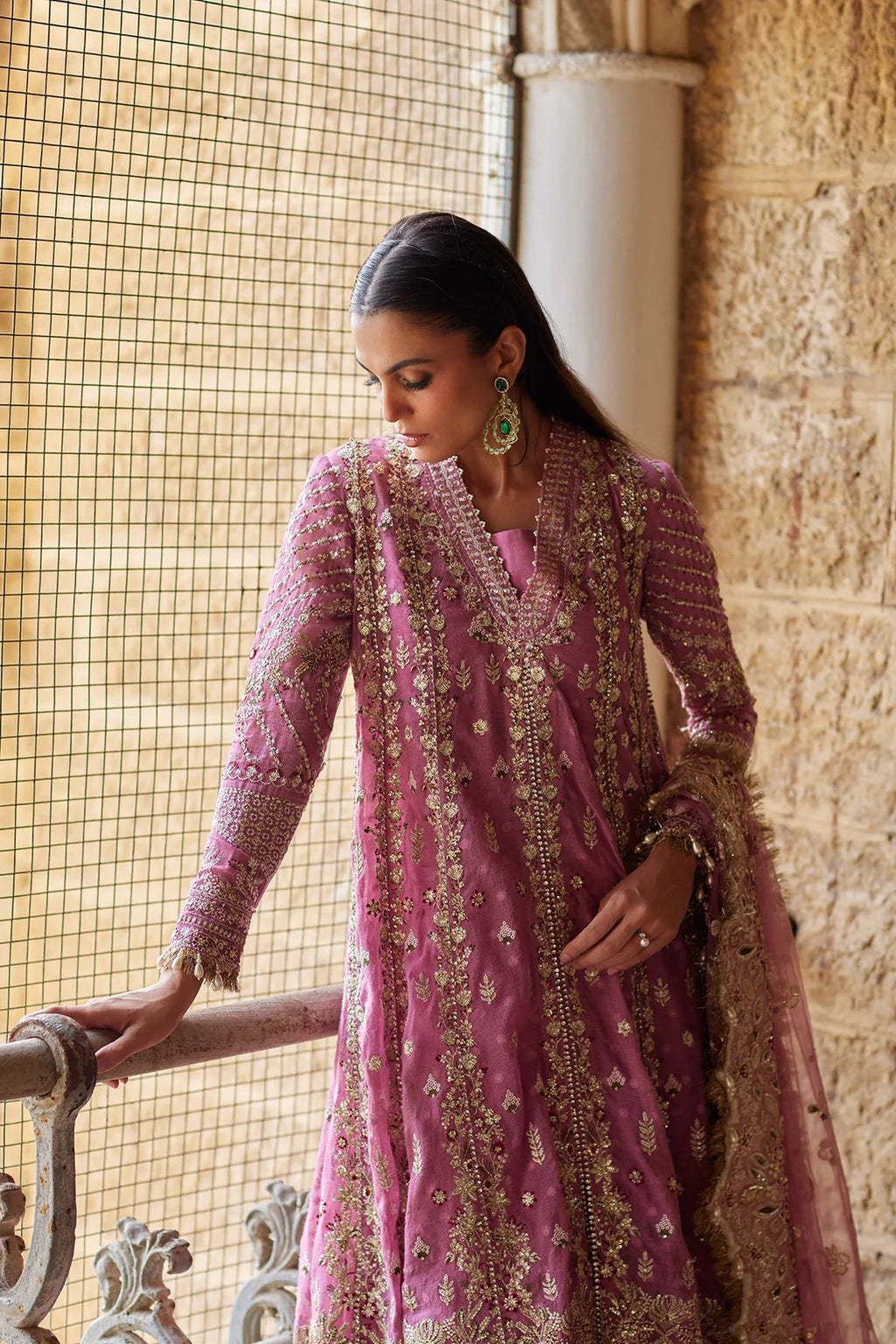 A woman wearing a pink traditional embroidered outfit with gold detailing, standing indoors near a stone wall and window screen.
