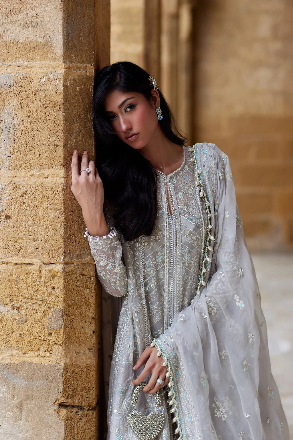 A woman wearing a heavily embellished light silver-gray traditional outfit with intricate embroidery and a matching sheer dupatta, posed against a warm beige stone column.