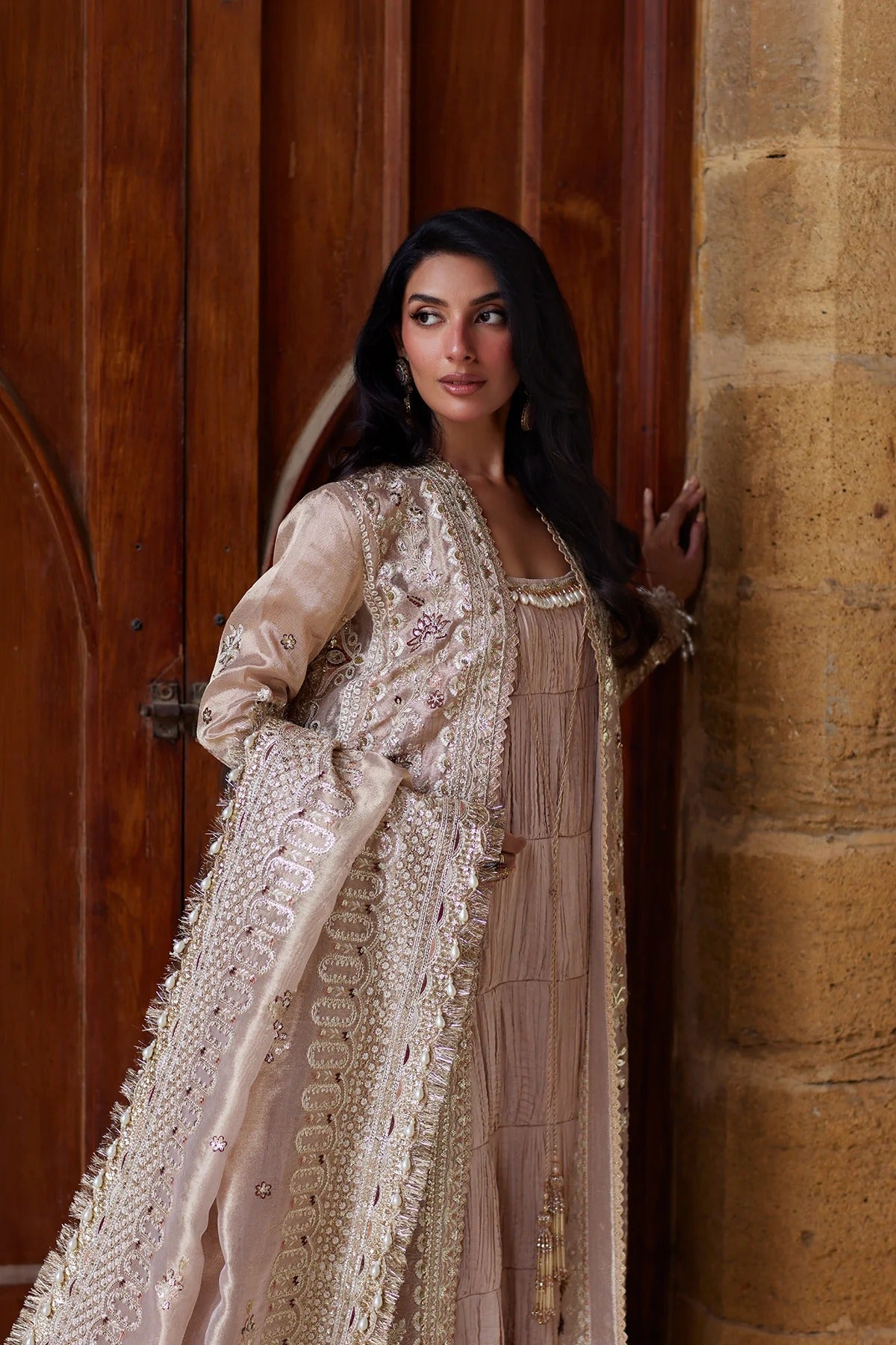 A woman models a traditional beige and gold embroidered outfit with a matching sheer dupatta, standing in front of a rustic wooden door and stone pillar.