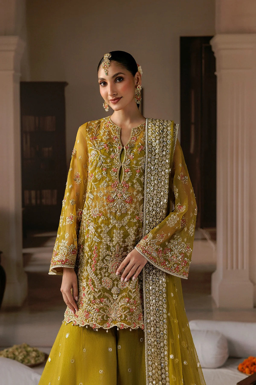 A woman models a traditional Indian salwar kameez suit in olive yellow with intricate gold, pink, and white embroidery. The set includes a heavily embroidered tunic (kameez) with a matching embroidered dupatta, paired with solid olive yellow palazzo pants.