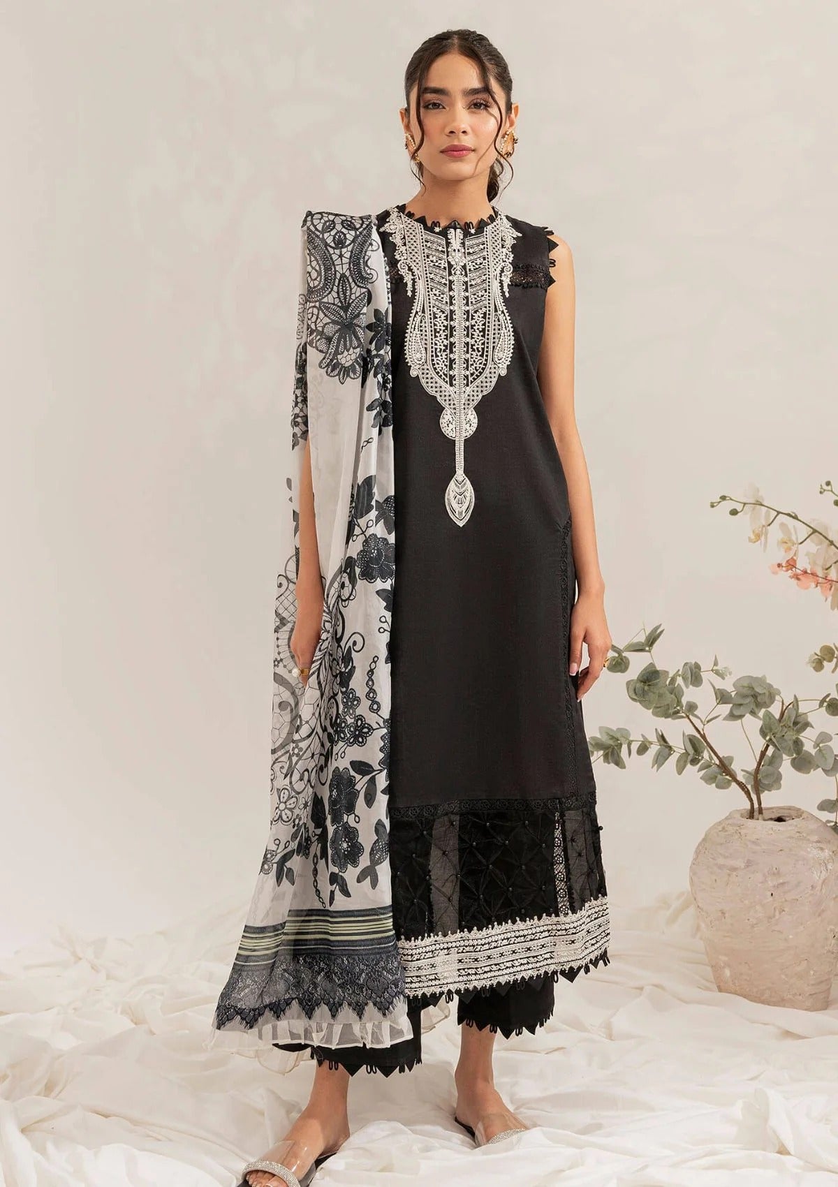 A woman models a black three-piece traditional outfit with intricate silver embroidery on the neckline, paired with a black and white patterned dupatta and black trousers, set against a neutral studio backdrop with a decorative plant in the background.