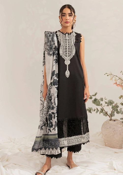 A woman models a black three-piece traditional outfit with intricate silver embroidery on the neckline, paired with a black and white patterned dupatta and black trousers, set against a neutral studio backdrop with a decorative plant in the background.