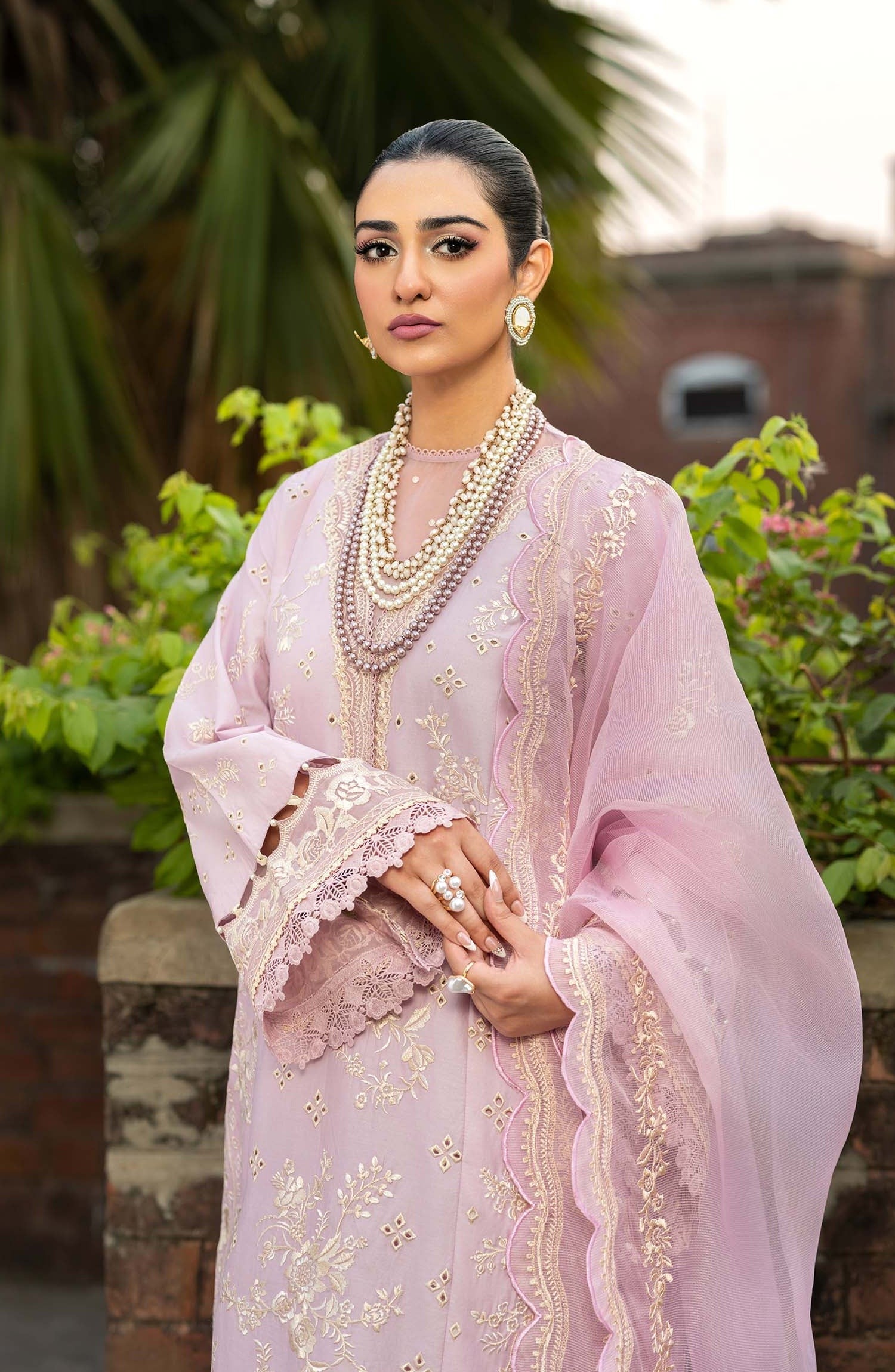 A woman wearing a light pink traditional outfit with gold embroidery and a matching sheer dupatta, styled with layered pearl necklaces and earrings, set against a soft-focus outdoor background of greenery and a stone wall.