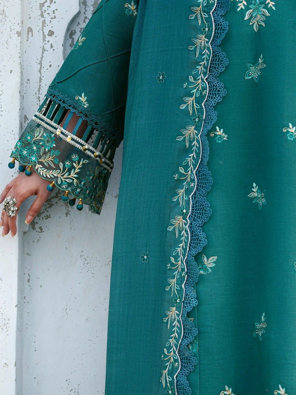A close-up of a teal green traditional outfit with intricate floral embroidery, scalloped lace borders, and decorative sleeve cuffs with tassel details, worn by a model whose hand is visible against a textured white wall.