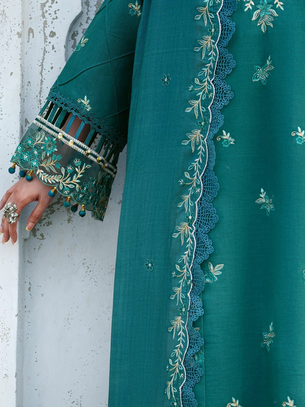 A close-up of a teal green traditional outfit with intricate floral embroidery, scalloped lace borders, and decorative sleeve cuffs with tassel details, worn by a model whose hand is visible against a textured white wall.