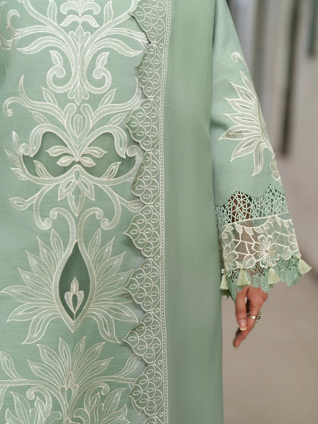 A close-up of a sage green traditional garment with intricate white embroidery and lace detailing on the sleeve and border.