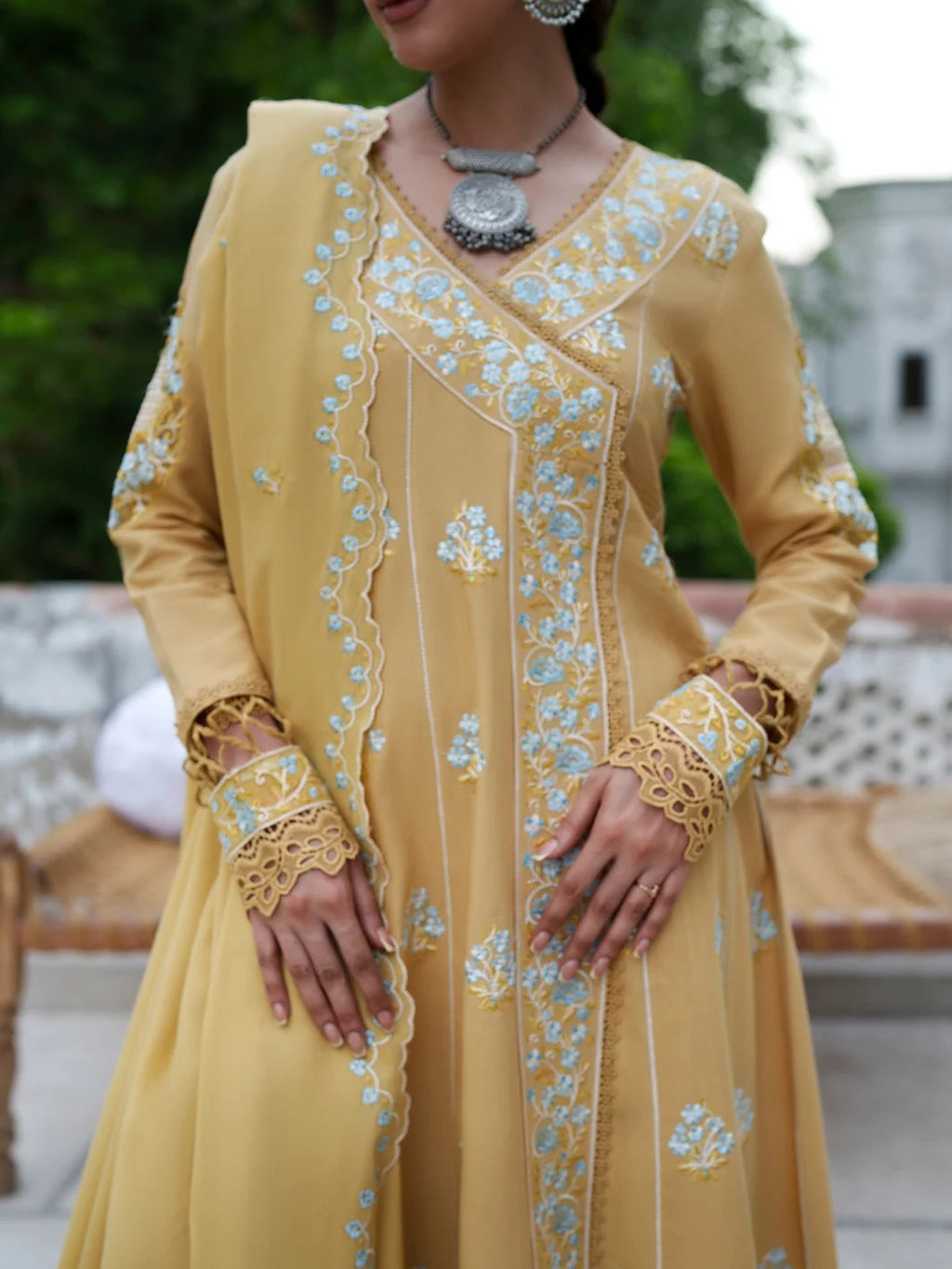 A woman wearing a long-sleeved mustard yellow traditional outfit with intricate light blue and white floral embroidery, lace cuffs, and a matching draped scarf, set against an outdoor background.
