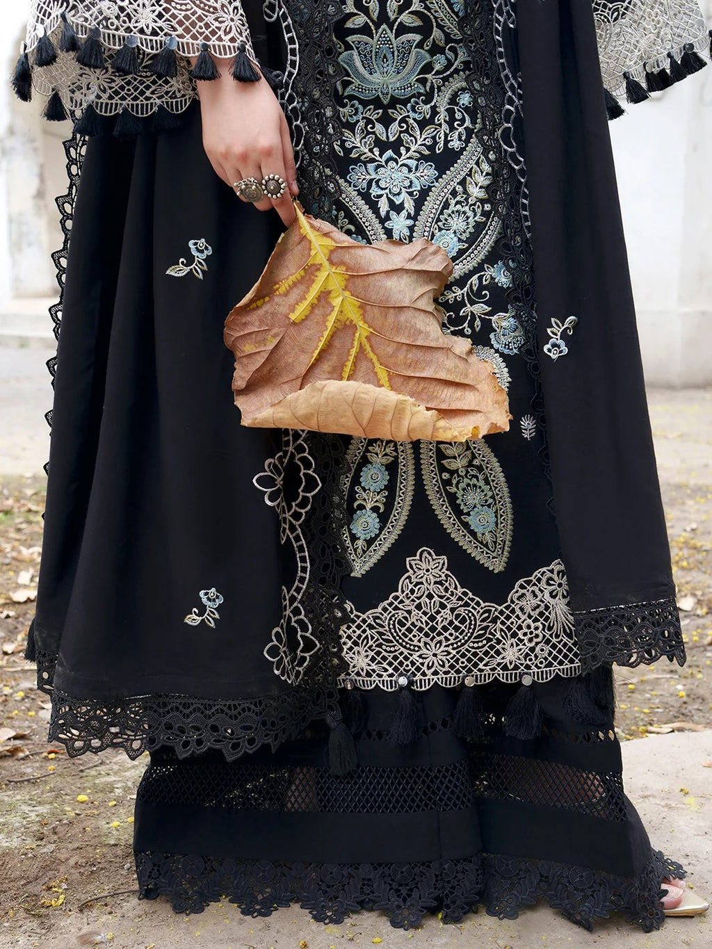 A close-up of a person wearing a black embroidered traditional outfit with intricate silver and blue floral embroidery, holding a dried brown leaf handbag with yellow-green veining.