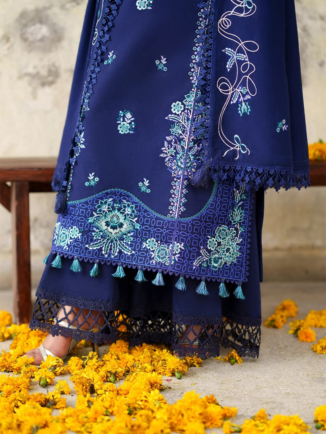 A close-up of a navy blue traditional outfit with intricate light blue and white embroidery, lace borders, and tassel details, worn by a model standing on a stone floor surrounded by yellow marigold flowers.