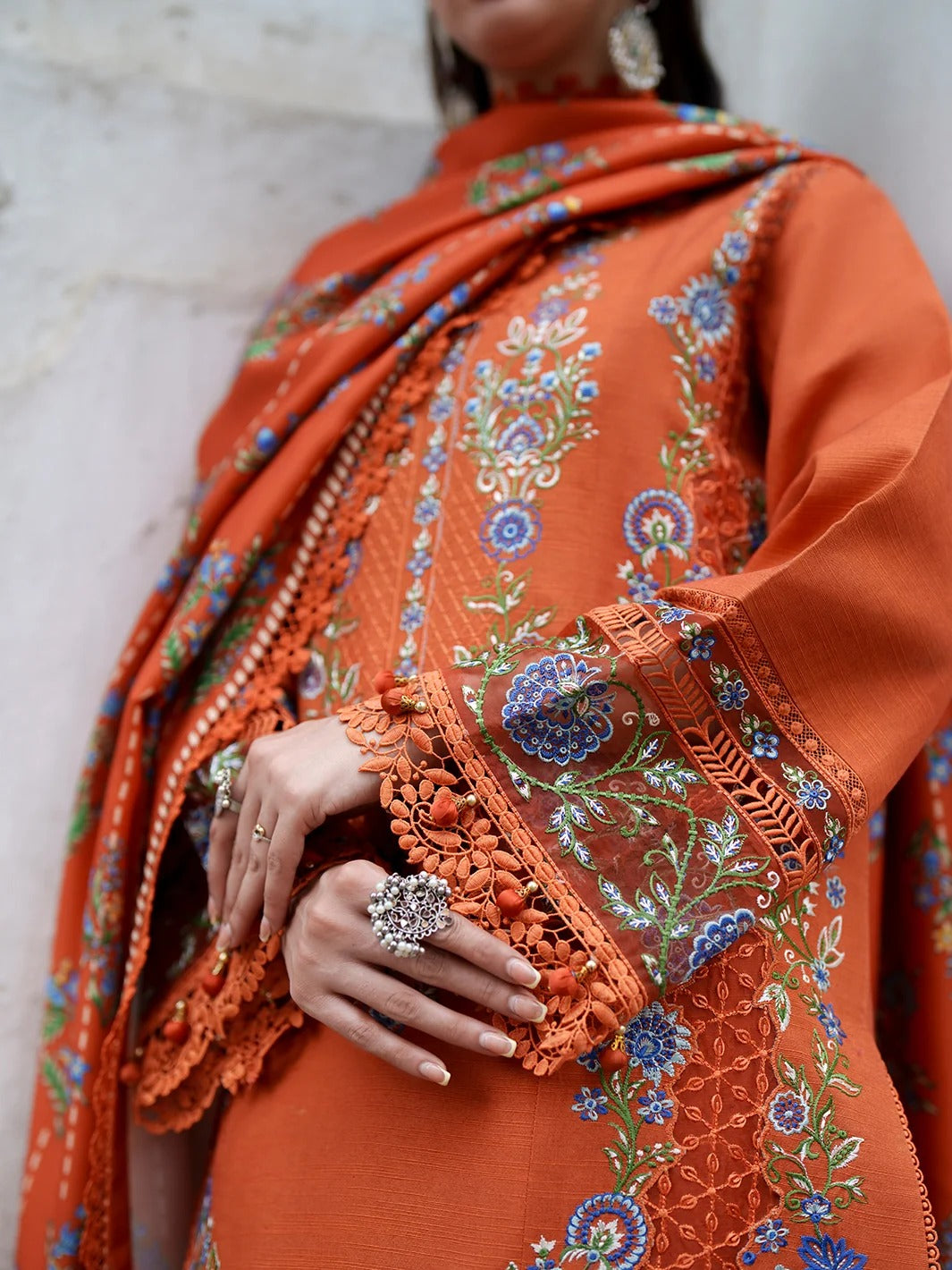 A close-up of a person wearing an orange embroidered traditional outfit with a matching draped scarf. The fabric features intricate floral embroidery in blue, green, and white, with detailed lace trim and tassels.