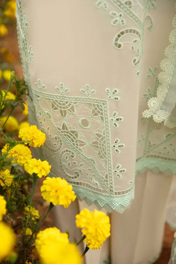 A close-up of a cream-colored eyelet lace garment with intricate mint green embroidery, styled with bright yellow flowers in the foreground.