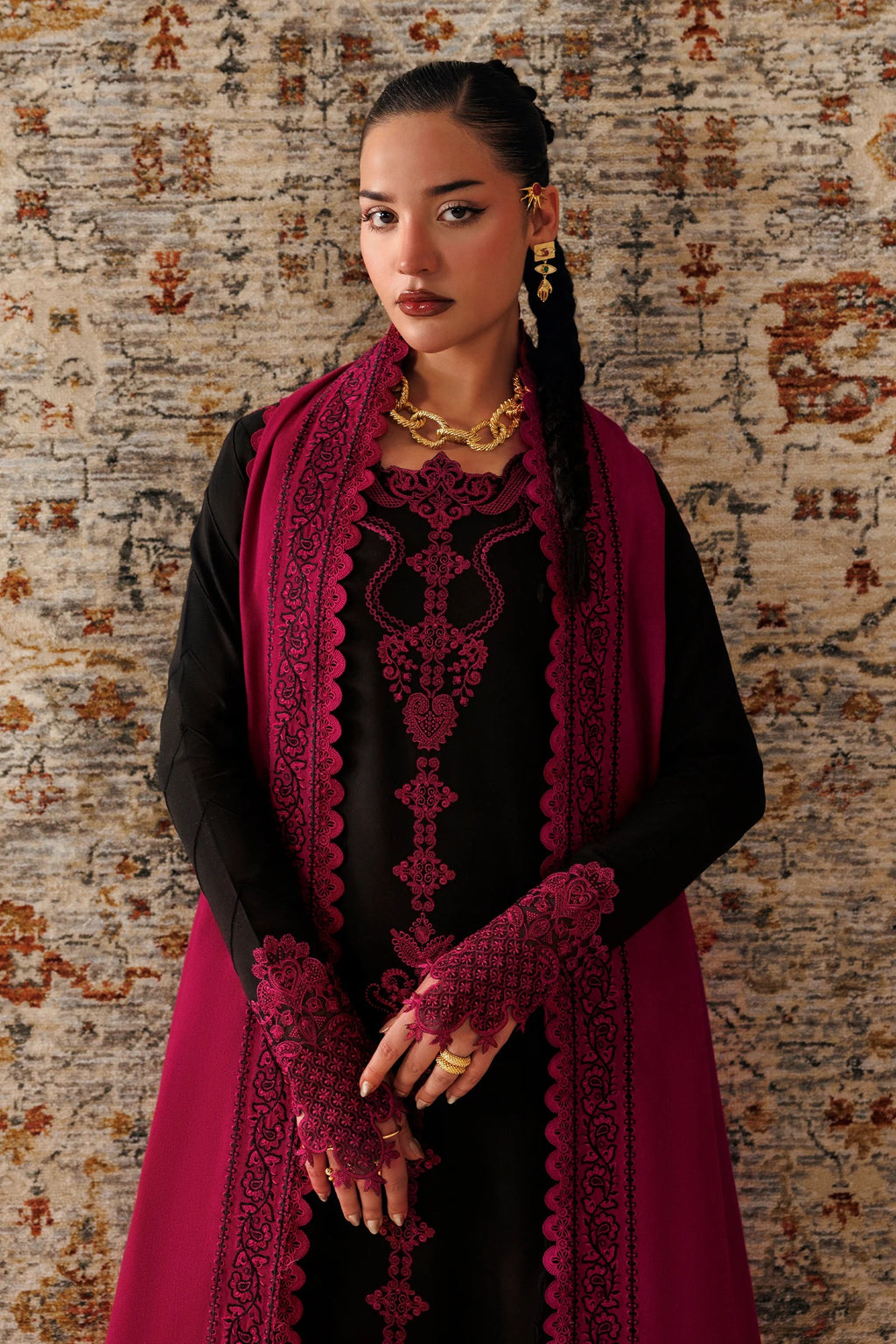 A woman models a traditional black dress with deep magenta embroidery and a matching magenta shawl, accessorized with gold jewelry, set against a textured patterned rug.