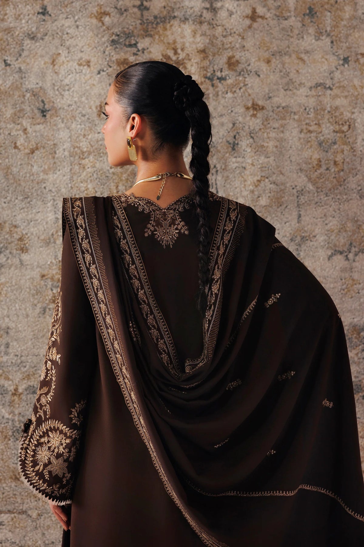 A woman shown from the back wearing a dark brown traditional outfit with intricate gold embroidery and a matching draped scarf, posed against a textured stone wall.