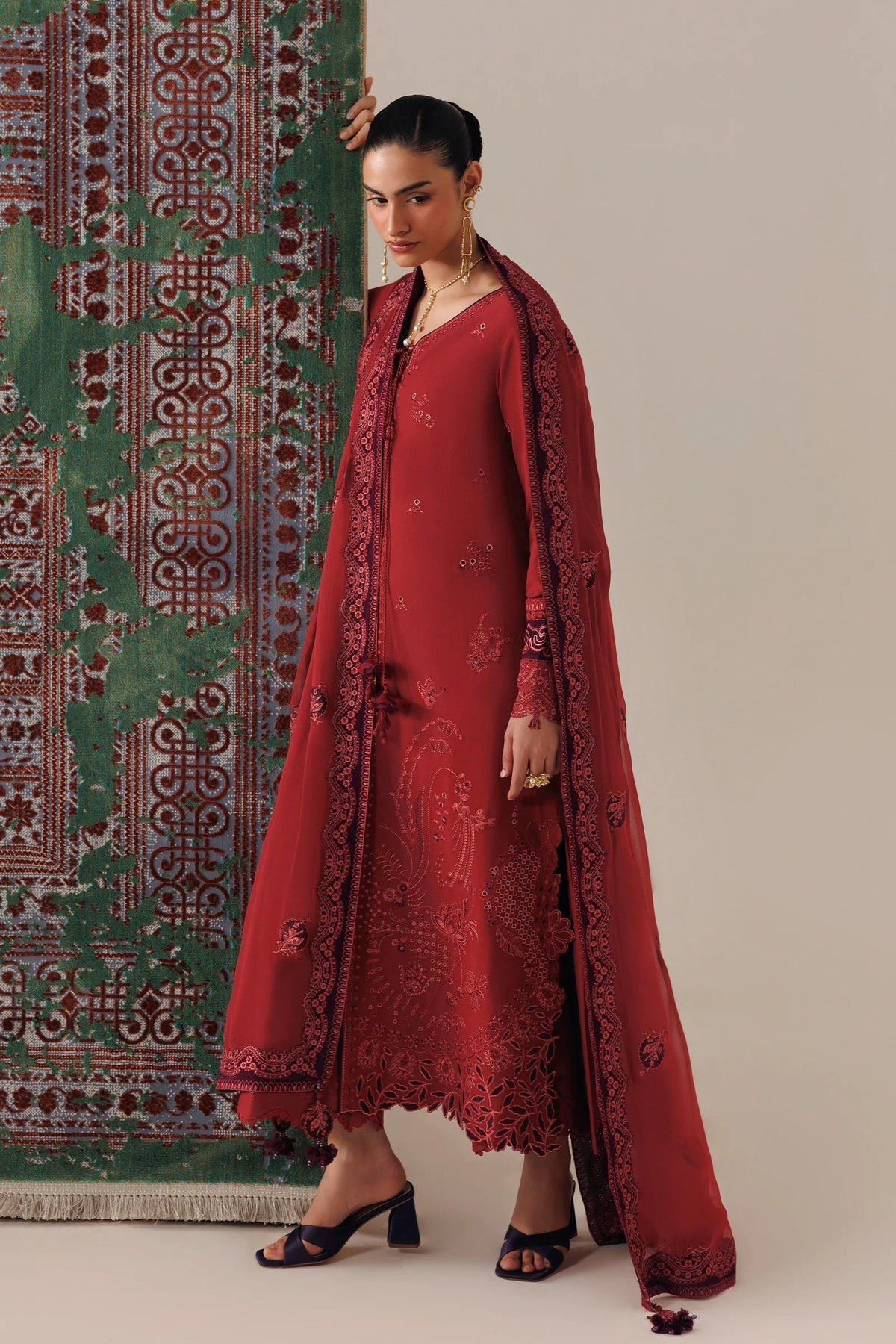 A woman models a traditional red salwar kameez set with an embroidered dupatta, standing in front of a beige wall and a green patterned rug.