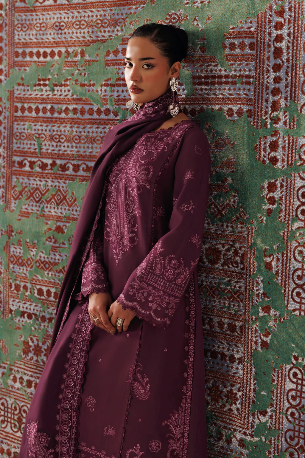 A woman wearing a deep plum purple embroidered traditional outfit with a matching scarf, standing against a patterned green and red wall.