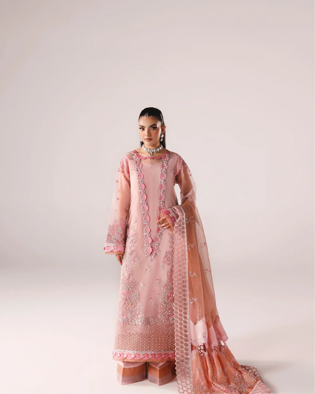 A model wearing a light pink traditional South Asian outfit with floral embroidery and a matching sheer dupatta, standing against a solid light beige studio background.