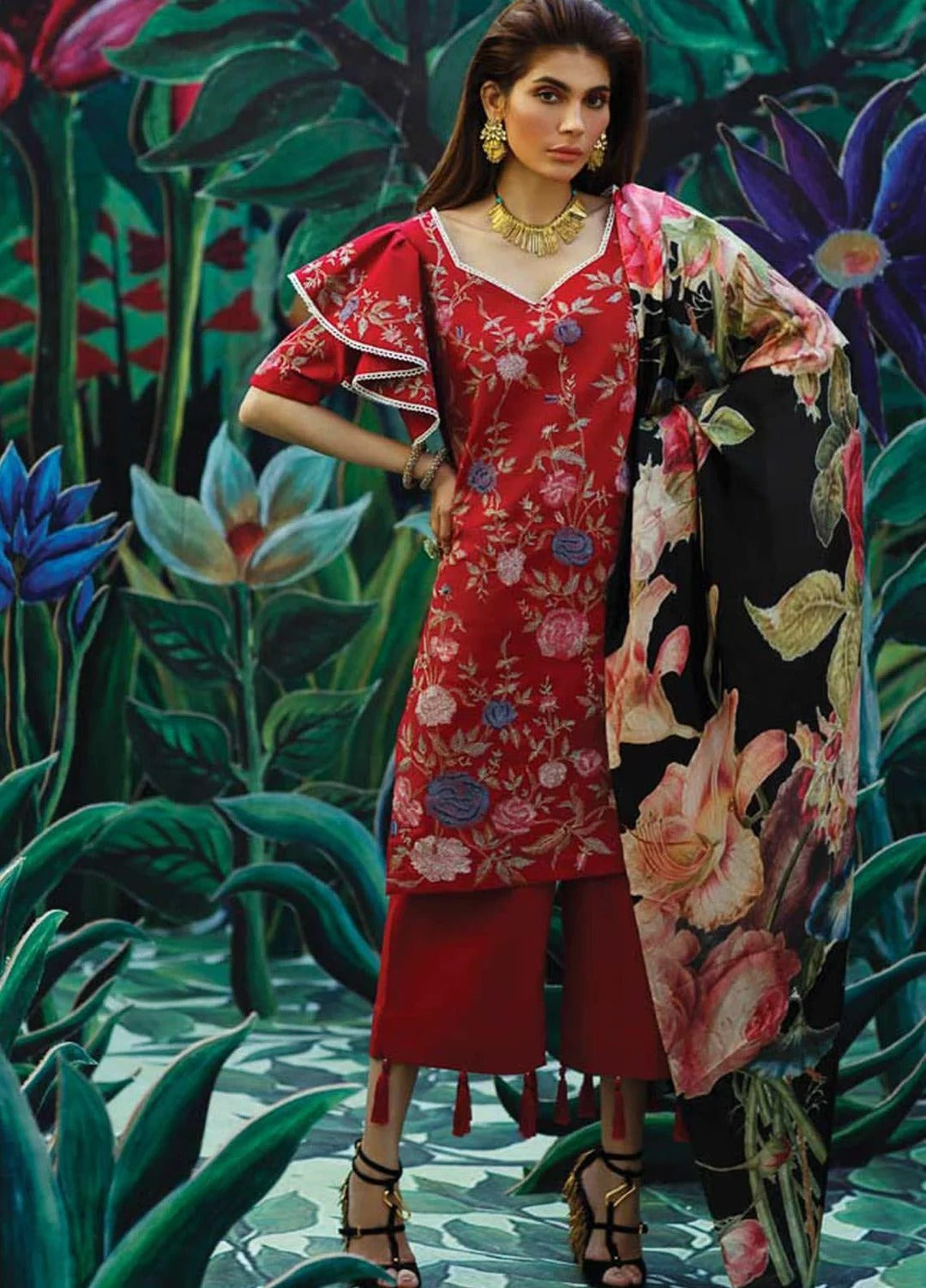 A woman models a red floral printed kurta with matching red pants and a black floral dupatta, posed against a painted tropical garden backdrop.