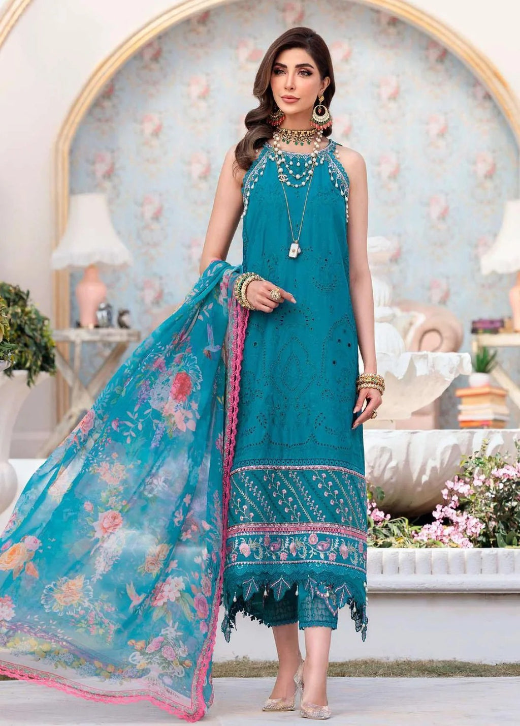 A woman models a teal blue ethnic three-piece suit set, featuring a sleeveless kurta with floral embroidery, matching cropped pants, and a sheer printed dupatta with pink borders, posed in front of a decorative archway background.