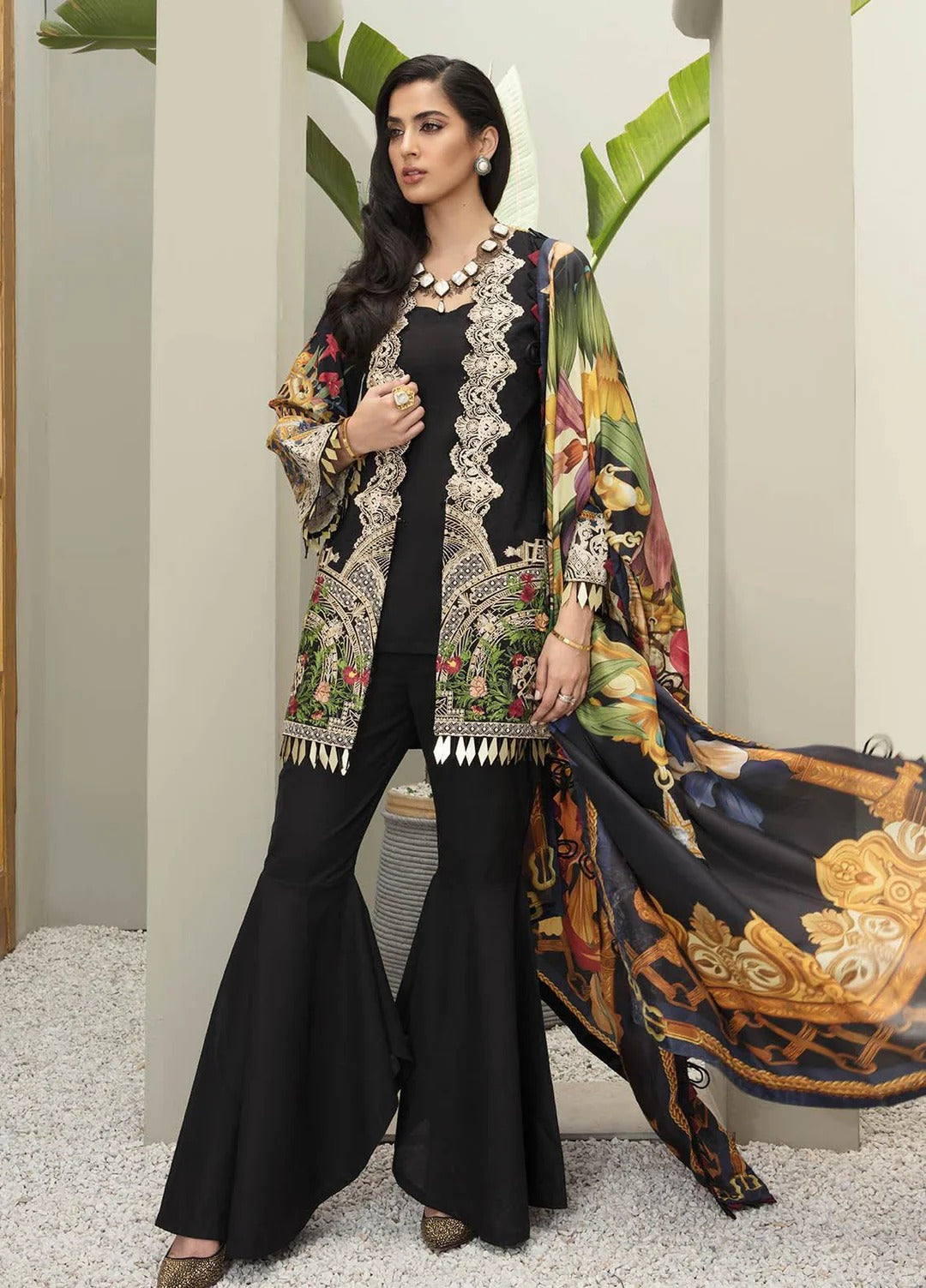 A woman models a black three-piece traditional outfit with ornate gold embroidery, flared pants, and a matching printed dupatta draped over one side, set against a neutral indoor backdrop with decorative plants.