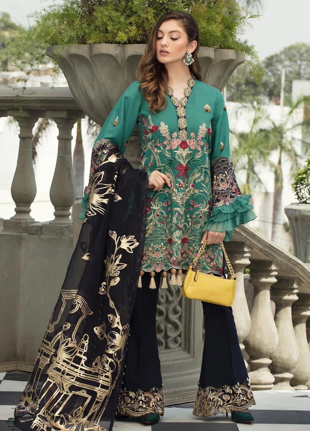 A woman models a three-piece traditional South Asian outfit. The set includes a teal green kurta with colorful floral embroidery, matching flared pants, and a black dupatta with gold patterns.