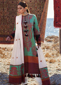 A woman models a traditional three-piece salwar kameez suit in mint green and maroon with intricate patterns, paired with a white dupatta featuring red motifs, set against a rustic background with patterned rugs and a rocky shoreline.