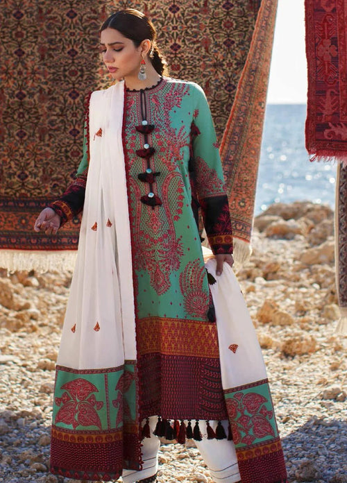 A woman models a traditional three-piece salwar kameez suit in mint green and maroon with intricate patterns, paired with a white dupatta featuring red motifs, set against a rustic background with patterned rugs and a rocky shoreline.