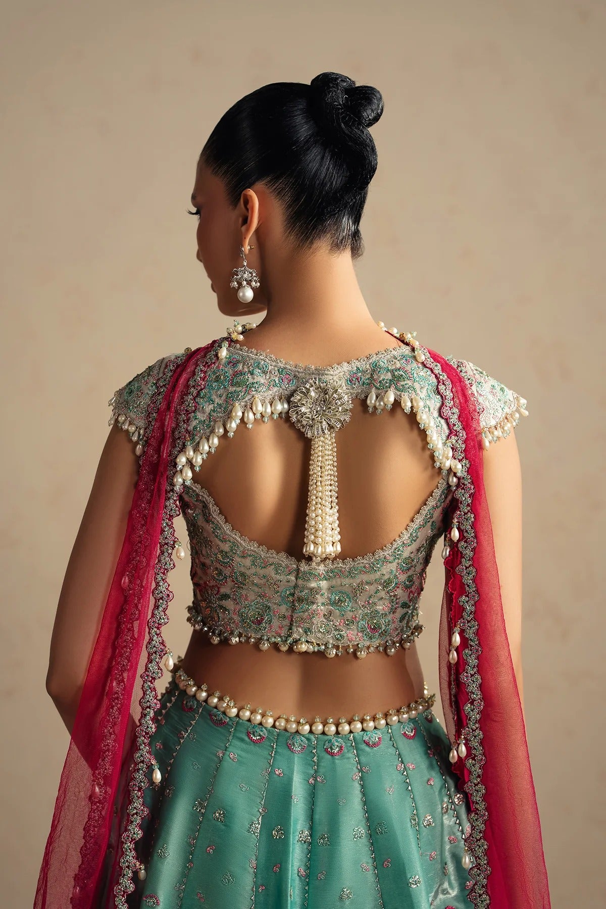 A woman shown from the back wearing an ornate teal embroidered lehenga choli set with a bright pink dupatta and heavy gold and pearl embellishments on the open back, against a plain beige studio background.