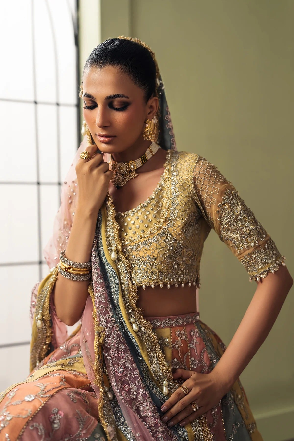 A woman models an ornate traditional Indian outfit featuring a heavily embroidered gold and silver blouse, a pink and gold lehenga skirt, and a matching dupatta, set against a softly lit indoor background.