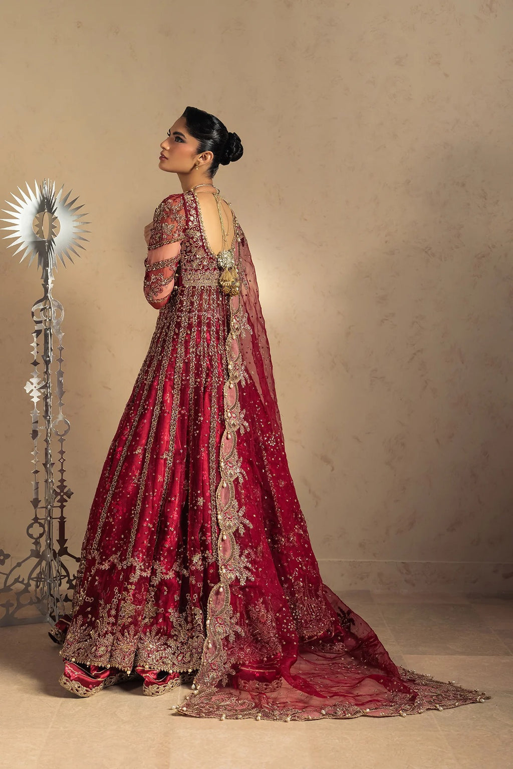 A woman wearing an ornate deep red and gold embroidered traditional gown stands in profile against a warm beige wall, with a decorative silver sunburst lamp on the left.