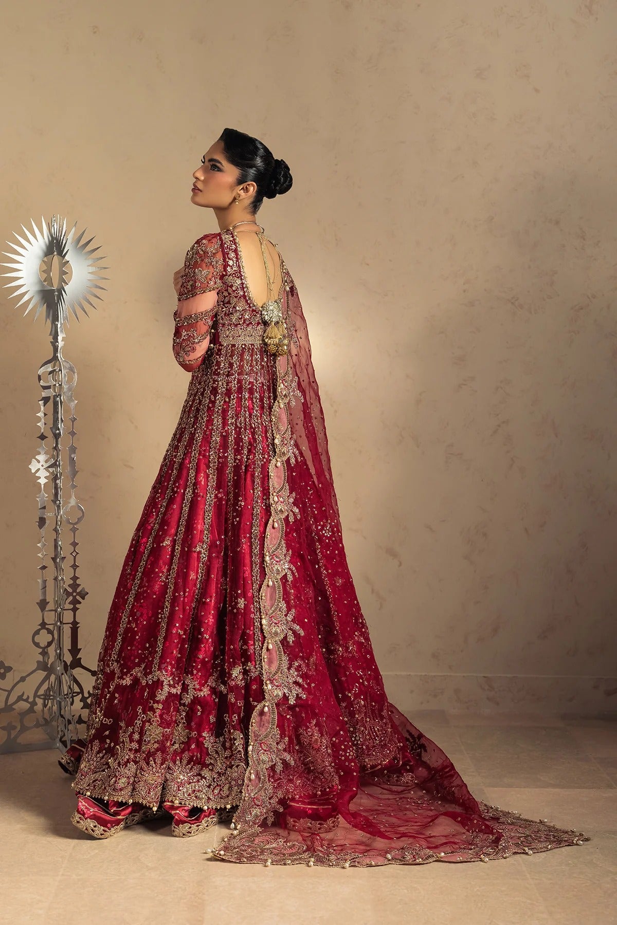 A woman wearing an ornate deep red and gold embroidered traditional gown stands in profile against a warm beige wall, with a decorative silver sunburst lamp on the left.
