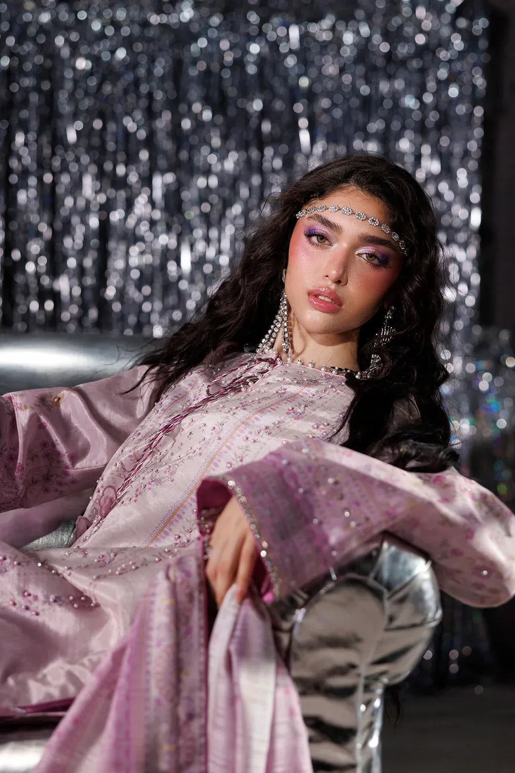 A woman with dramatic eye makeup and a jeweled headpiece reclines on a metallic chair in front of a sparkling silver curtain backdrop, wearing a lavender satin embroidered traditional outfit with wide sleeves.