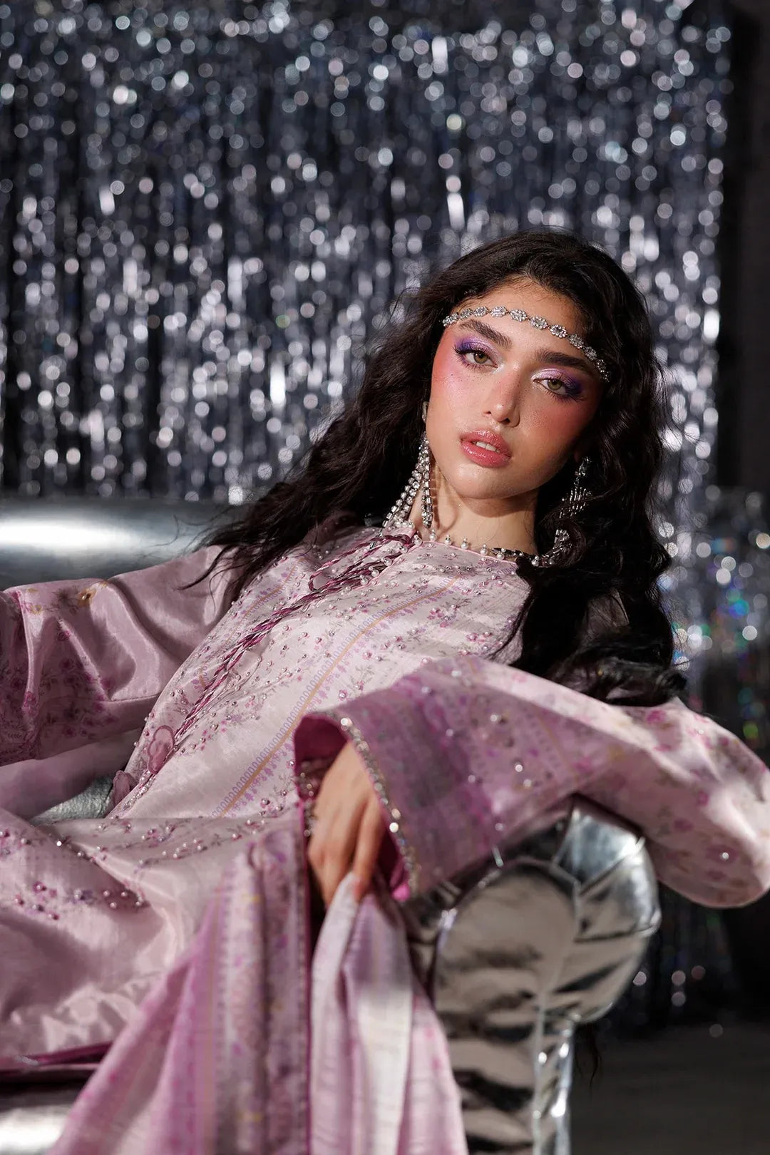 A woman with dramatic eye makeup and a jeweled headpiece reclines on a metallic chair in front of a sparkling silver curtain backdrop, wearing a lavender satin embroidered traditional outfit with wide sleeves.