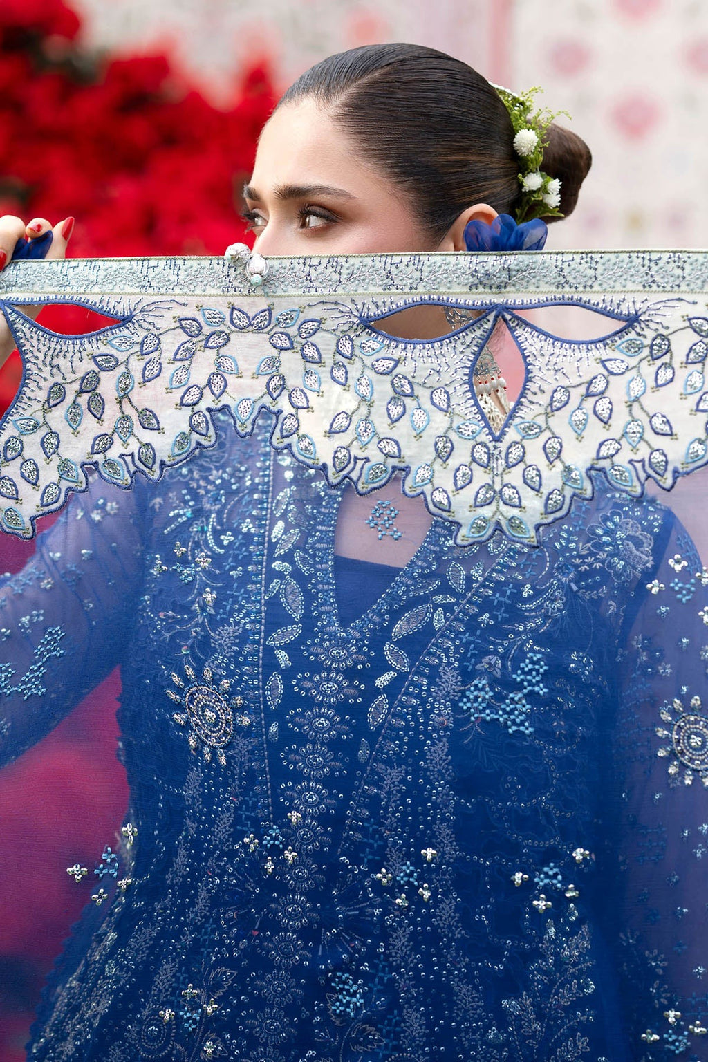 A woman holds up a detailed view of a royal blue embroidered traditional garment, showing off its intricate silver and white floral embroidery and scalloped collar, with a blurred red floral background.