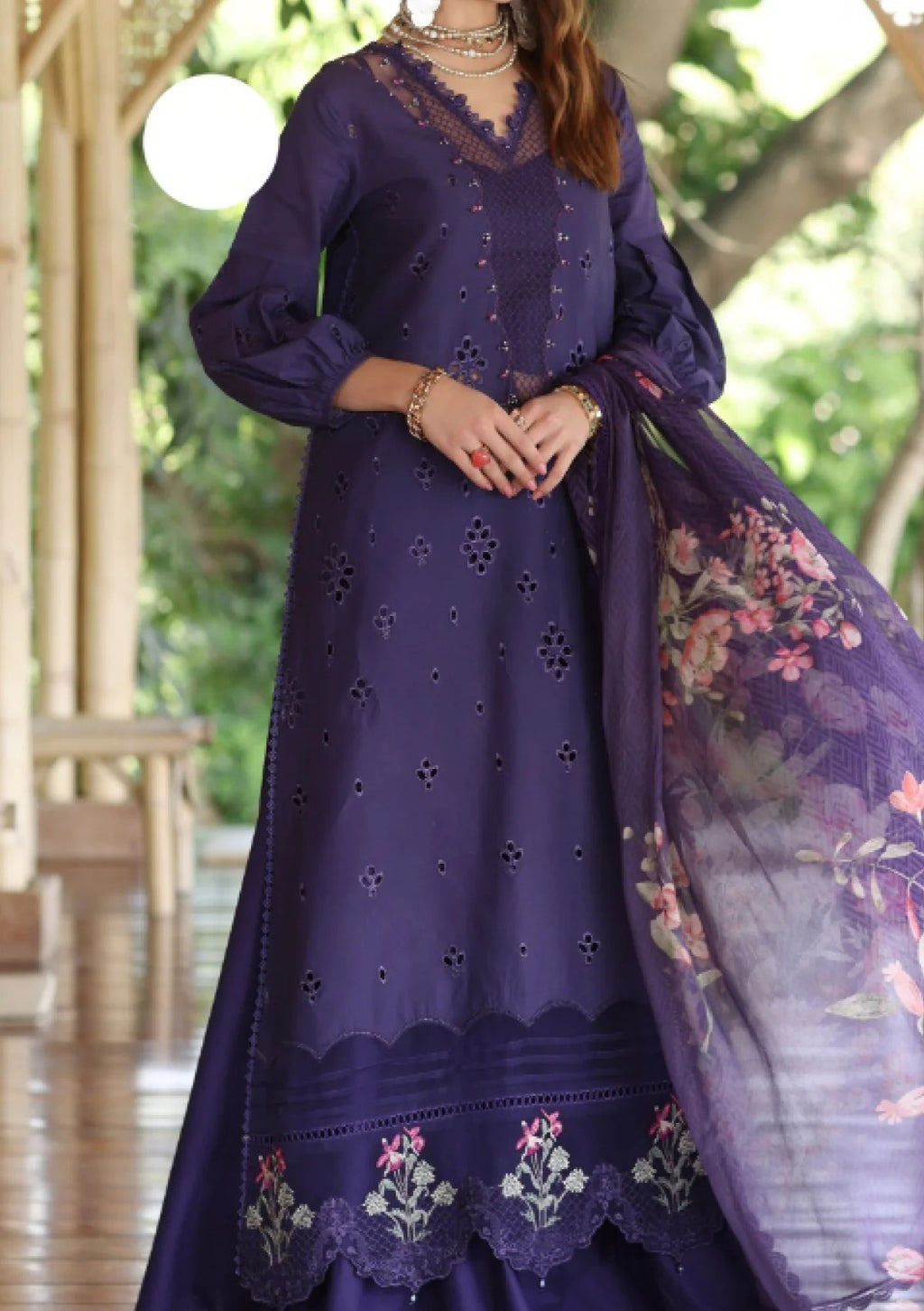 A woman wearing a deep purple traditional three-piece outfit with floral printed dupatta, lace-trimmed neckline, and embroidered floral border at the hem, photographed in a softly blurred outdoor corridor.