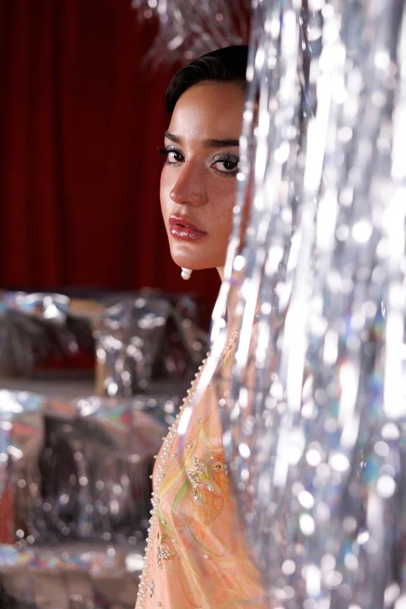 A woman in a peach embellished dress peering from behind a shiny silver tinsel curtain, with a red curtain backdrop and blurred silver wrapped items in the background.