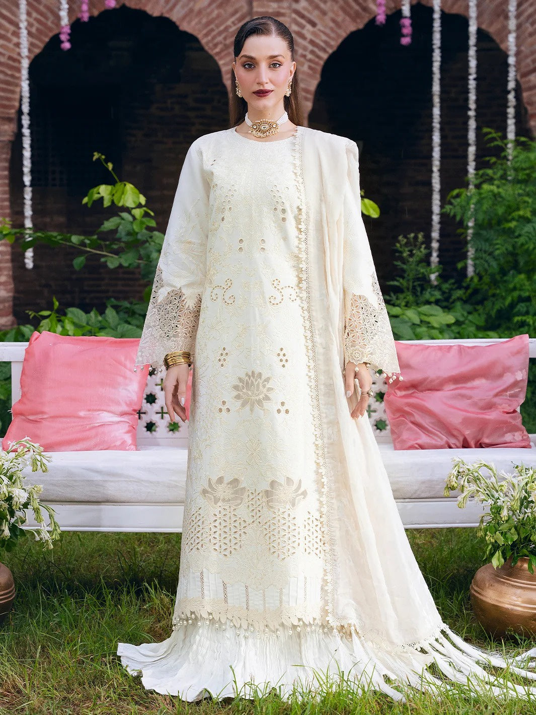 A woman stands outdoors wearing a long cream-colored embroidered traditional outfit with a matching draped scarf. The outfit features intricate floral embroidery and cutwork details.