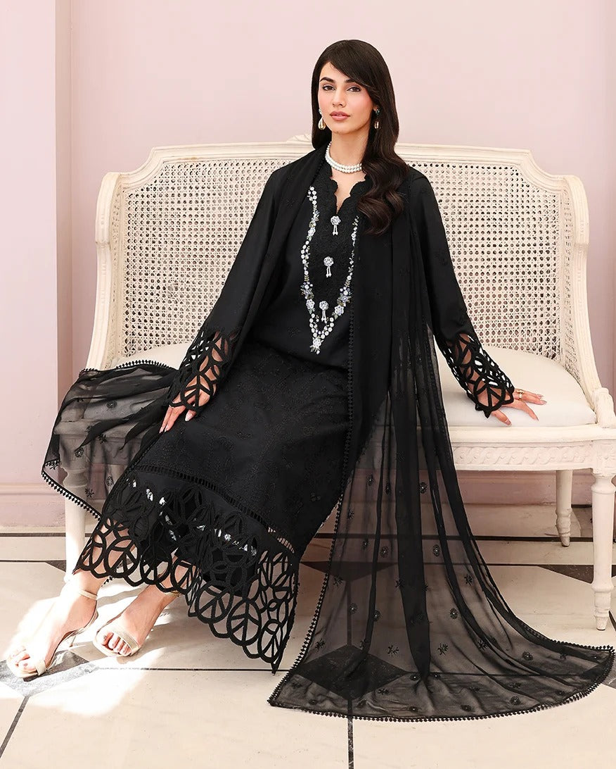 A woman models a black embroidered traditional outfit with a matching sheer draped overlay and long sleeves, seated on a white woven bench against a pale pink wall.
