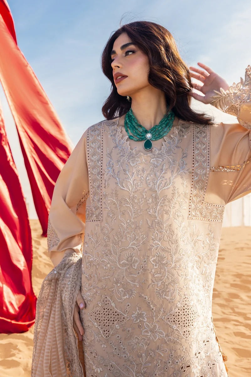A woman wearing a beige embroidered traditional outfit with a matching dupatta and a green statement necklace, standing in a sandy desert setting with a red fabric drape in the background.