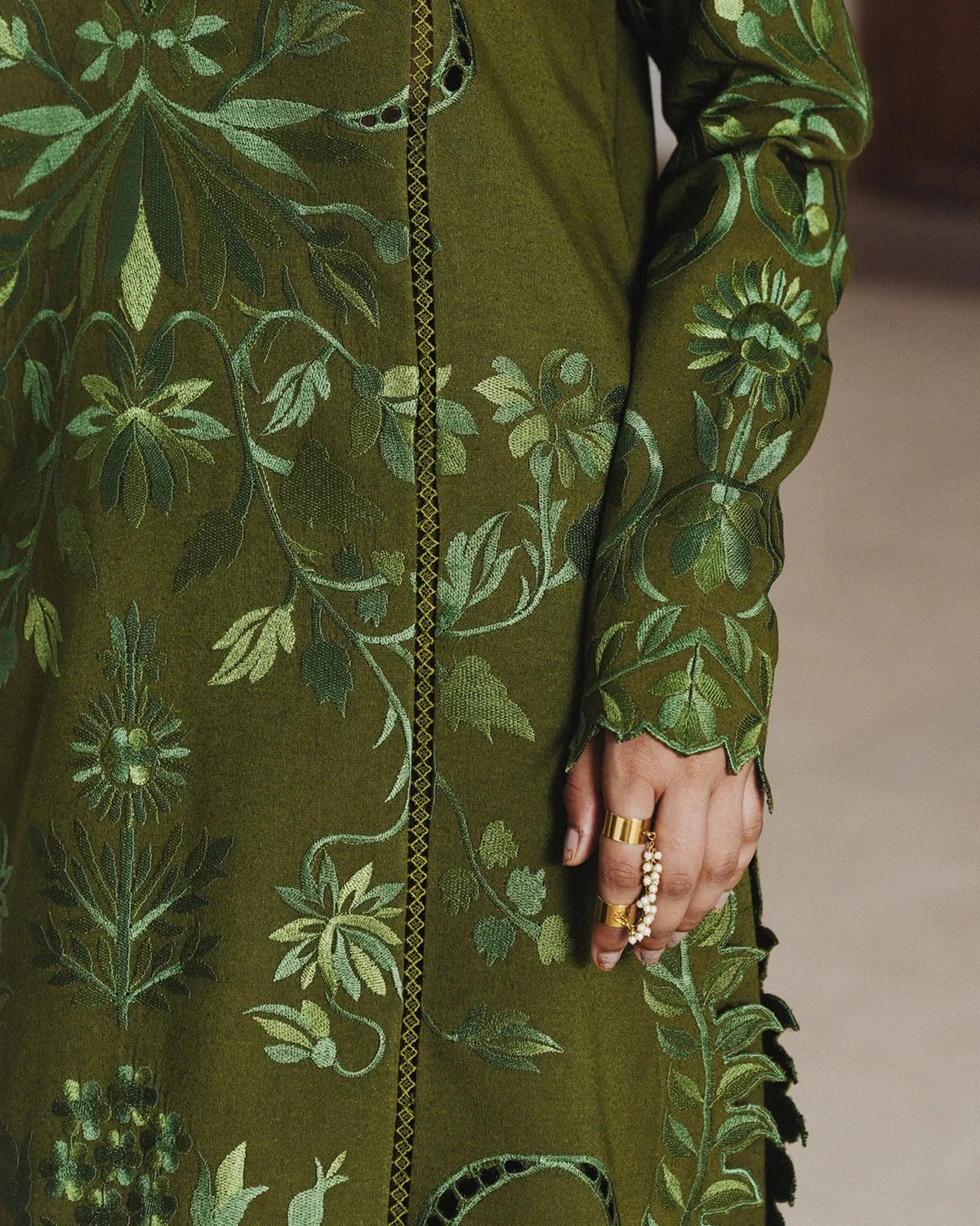 Close-up of an olive green embroidered kurta featuring intricate floral and vine patterns in shades of green, with a hand adorned with gold rings visible at the side.